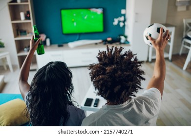 African American Sports Fan Couple Watching Important Soccer Match On TV While Drinking Beer And Cheering At Home. Guy Holding A Soccer Ball. Cheering For A Favorite Team. Rear View.