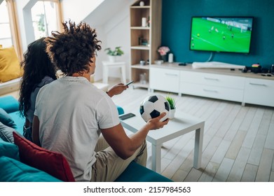 African American Sports Fan Couple Watching Important Soccer Match On TV While Drinking Beer And Cheering At Home. Guy Holding A Soccer Ball. Cheering For A Favorite Team.