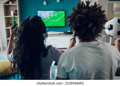 African American Sports Fan Couple Watching Important Soccer Match On TV While Drinking Beer And Cheering At Home. Guy Holding A Soccer Ball. Cheering For A Favorite Team. Rear View.