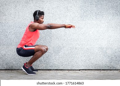 african american sports guy working out with headphones outdoors against wall, athletic man crouches and does physical exercises, copy space