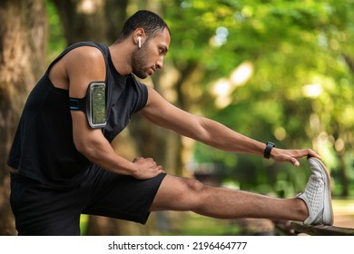 African American Sportsman Exercising Outdoors At Public Park, Young Black Guy In Sportswear With Arm Pocket For Cell Phone And Wireless Earbuds Stretching His Body, Side View, Copy Space