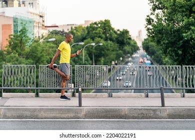 African American sportsman stretching his right leg and holding phone in the footbridge at day. - Powered by Shutterstock