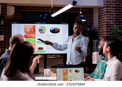 African American Startup Employee Holding Remote Control Presenting Sales Pie Charts On Tv Screen In Night Meeting. Woman Doing Overtime At Work Talking About Business Strategy With Diverse Team.