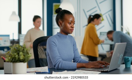 African American Startup Employee Sitting At Desk Opening Laptop And Starting To Type On Keyboard Working On Sales Statistics. Business Woman In Modern Busy Office Beginning Work In The Morning.