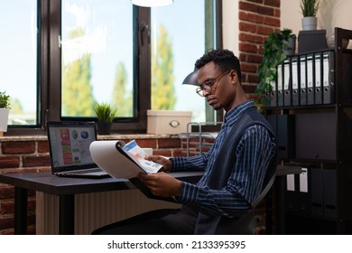 African American Startup Owner With Glasses Holding Clipboard And Paper With Bussiness Charts Comparing Data With Sales Results On Laptop. Entrepreneur Analyzing Marketing Indicators On Laptop.