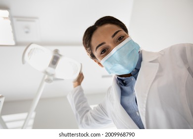 African American Stomatologist Lady Adjusting Medical Lamp Before Check Up With Patient, Professional Black Dentist Doctor Woman Wearing Protective Medical Mask Ready For Dental Treatment, Low Angle - Powered by Shutterstock