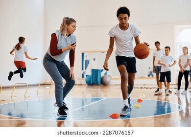 African American student practicing basketball while having PE class with classmates and sports teacher at school gym.  - Powered by Shutterstock