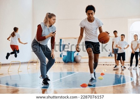 African American student practicing basketball while having PE class with classmates and sports teacher at school gym. 