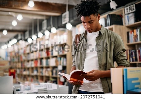 African American student reading a book while standing at bookstore.
