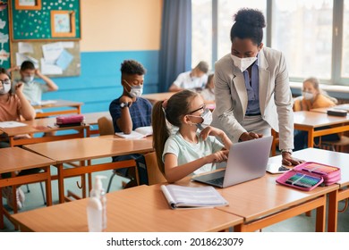 African American teacher assisting schoolgirl in using computer during a class in the classroom. They are wearing face masks due to coronavirus pandemic.   - Powered by Shutterstock