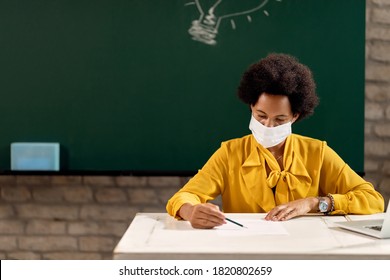 African American Teacher Wearing Protective Face Mask While Reading Students Tests In The Classroom. 