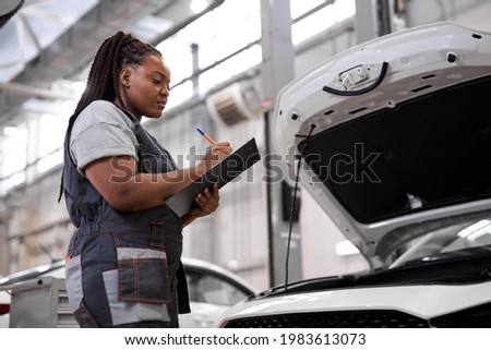 African american technician female concentrated to check list for maintenance in car garage service, wearing overalls uniform, looking focused while writing in documents tablet. side view