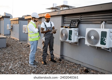 African american technician with tablet in hands showing fixed air conditioner to caucasian manager in white hard hat on rooftop. Factory workers cooperating for maintaining modern equipment. - Powered by Shutterstock