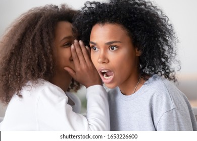 African american teen daughter whispering in ear of shocked young mother telling secret rumors or unbelievable news, surprised black mom sister friend and teenager girl talking gossiping at home - Powered by Shutterstock