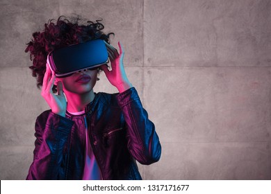 African American Teen Girl In Trendy Outfit Wearing VR Goggles And Looking Away While Standing Near Concrete Wall