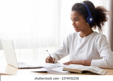 African American teen girl wearing headphones learning language online, using laptop, looking at screen, doing school tasks at home, writing notes, listening to lecture or music, distance education
