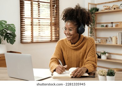 African American teen girl wearing headphones learning language online, using laptop, looking at screen, doing school tasks at home, writing notes, listening to lecture or music, distance education - Powered by Shutterstock