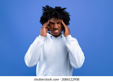 African American teen guy suffering from severe headache, massaging temples over blue studio background. Black youth having hypertension or migraine, feeling stressed or desperate