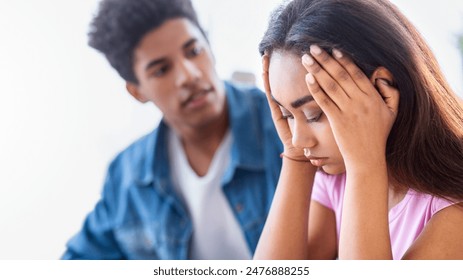 African American teen guy, wearing a blue denim shirt, looks at a teen girl with concern. The girl is seated with her hands covering her face, expressing sadness and distress. - Powered by Shutterstock