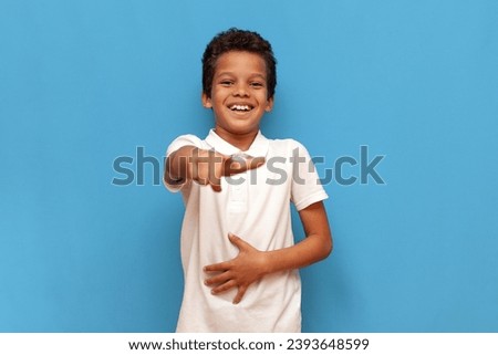 african american teenage boy in white polo taunts and jokes and points with hand forward on blue isolated background, cheerful child laughs and makes fun