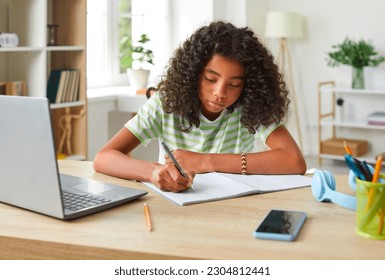 African American teenage girl handwriting in notebook while doing homework using laptop. Concentrated cute curly girl sitting at table in her room and studying remotely. Homeschooling concept. - Shutterstock ID 2304812441