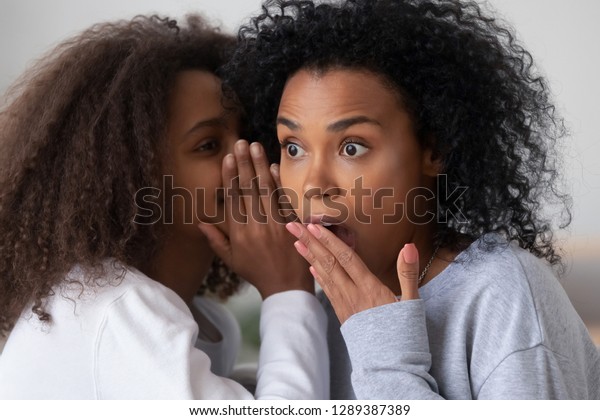 African American teenage girl tell secret to\
surprised young mom or nanny, teen daughter share gossip whispering\
in ear to shocked black mother, young parent and kid spend time\
together talking