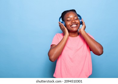 African American Teenager Listening Music Using Headphones Dancing Alone Enjoying Joyful Playlist In Studio With Blue Background. Portrait Of Smiling Young Woman Having Fun On Song Rhythm