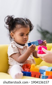 African American Toddler Girl Sitting On Couch And Playing Building Blocks With Mother On Blurred Foreground