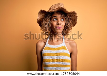African american tourist woman with curly on vacation wearing summer hat and striped t-shirt smiling looking to the side and staring away thinking.