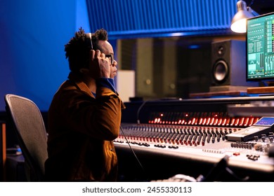 African american tracking engineer processing and editing music in studio control room, operating mixing console with moving sliders and colored meters. Audio technician deals with stereo gear.