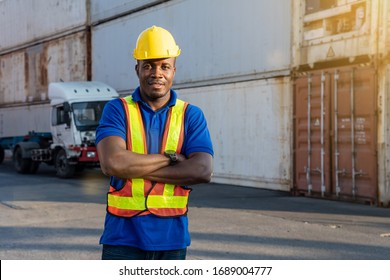 African American Transportation Factory Truck Driver With Yellow Helmet And Safety Vest Is Standing And Smiling By Action Arms Crossed In Front Of Lorry At Container Yard Of Port On Evening.