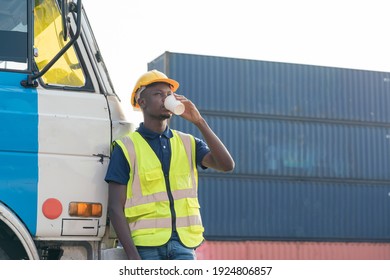 African American Transportation Factory Truck Driver With Helmet And Safety Vest Is Standing And Drink Cup Of Coffee At Heavy Lorry Of Container Yard In Logistic Construction Port. Black Skin Worker.