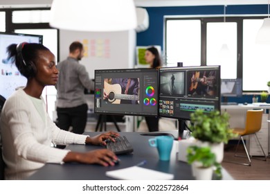 African american video editor wearing headset editing movie using post production software working in start up company office. Black creator processing video footage on computer with dual displays