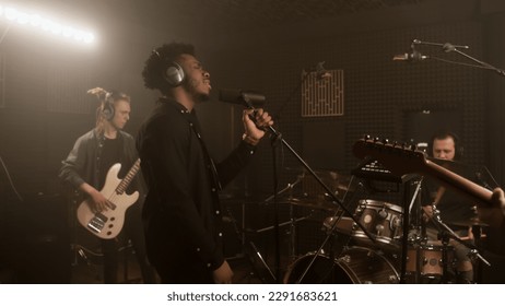 African American vocalist keeps hands on professional microphone stand and sings new composition in sound recording studio. Band plays on musical instruments at background. Music production concept. - Powered by Shutterstock