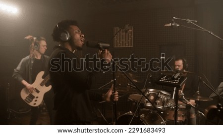 African American vocalist keeps hands on professional microphone stand and sings new composition in sound recording studio. Band plays on musical instruments at background. Music production concept.