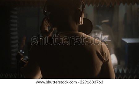African American vocalist sings composition into microphone in soundproof room. Experienced audio engineer or producer works with singer in sound recording studio. Music production concept. Back view.