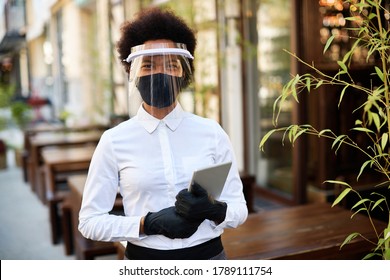 African American waitress wearing protective face mask and visor while holding touchpad and reopening sidewalk cafe during COVID-19 epidemic. - Powered by Shutterstock