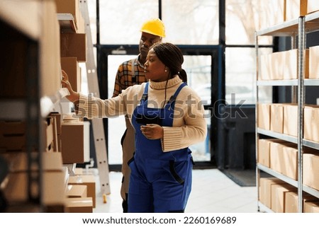 African american warehouse operatives standing in aisle near shelf full of cardboard boxes. Delivery service storehouse manager searching customer order and taking parcel package