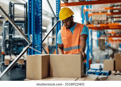 African american warehouse worker is seen handling boxes in a large storage area, equipped with shelves and racks, the worker is dressed in a hard hat and safety vest, ensuring orderliness. - Powered by Shutterstock