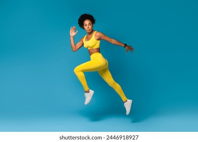 African American woman in action wearing vibrant yellow sports attire performing a jumping pose on a blue background - Powered by Shutterstock