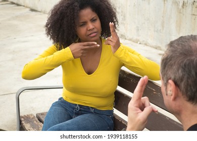 African American Woman With Afro Hair And Yellow T-shirt Speaking Sing Language Sitting On A Wooden Bench.