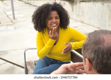 African American Woman With Afro Hair And Yellow T-shirt Speaking Sing Language Sitting On A Wooden Bench.