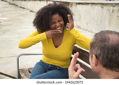 African American Woman With Afro Hair And Yellow T-shirt Speaking Sing Language Sitting On A Wooden Bench.