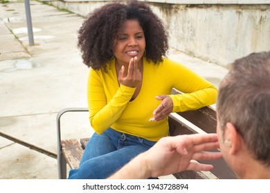 African American Woman With Afro Hair And Yellow T-shirt Speaking Sing Language Sitting On A Wooden Bench.