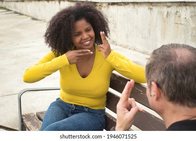 African American Woman With Afro Hair And Yellow T-shirt Speaking Sing Language Sitting On A Wooden Bench.