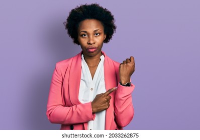 African american woman with afro hair wearing business jacket in hurry pointing to watch time, impatience, looking at the camera with relaxed expression