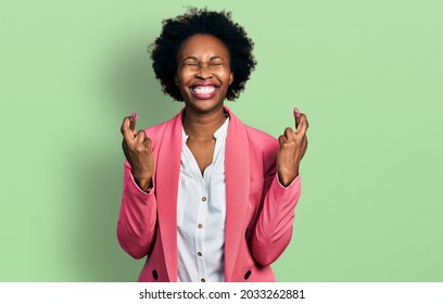 African american woman with afro hair wearing business jacket gesturing finger crossed smiling with hope and eyes closed. luck and superstitious concept. 