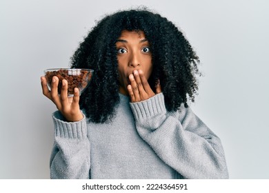 African American Woman With Afro Hair Holding Raisins In Bowl Covering Mouth With Hand, Shocked And Afraid For Mistake. Surprised Expression 