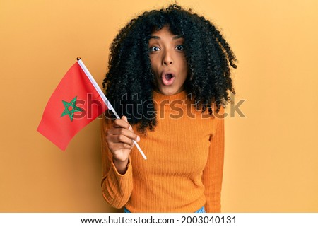 African american woman with afro hair holding morocco flag scared and amazed with open mouth for surprise, disbelief face 