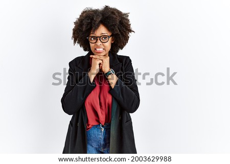African american woman with afro hair wearing business jacket and glasses laughing nervous and excited with hands on chin looking to the side 